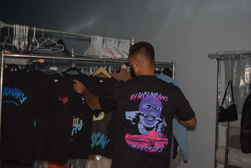 Person wearing a black t-shirt with colorful graphics and text, standing in front of a rack of clothes.
